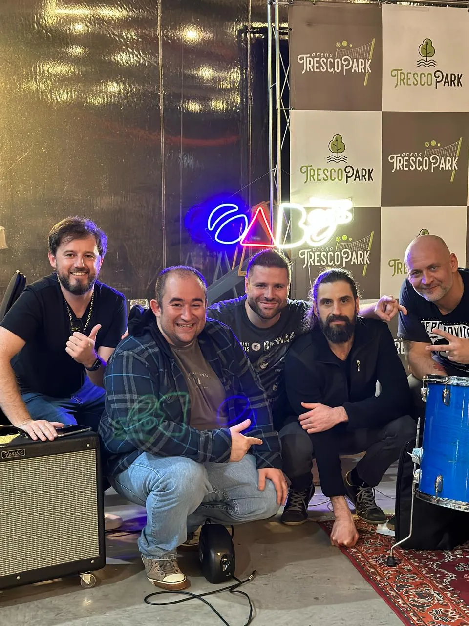 Integrantes da OABoys sorrindo ao lado do letreiro neon da banda na Arena Tresco Park