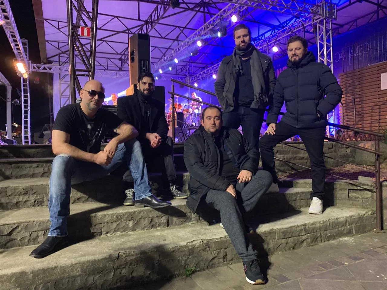 Integrantes da OABoys nas escadarias ao lado de um palco iluminado, à noite
